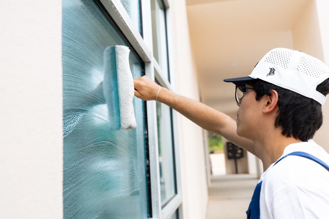 Person cleaning a window with a roller, wearing a cap and glasses, in a bright environment.