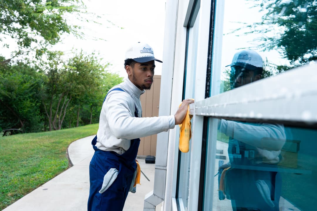 Window cleaner using a microfiber cloth on glass, wearing a cap and uniform outdoors.
