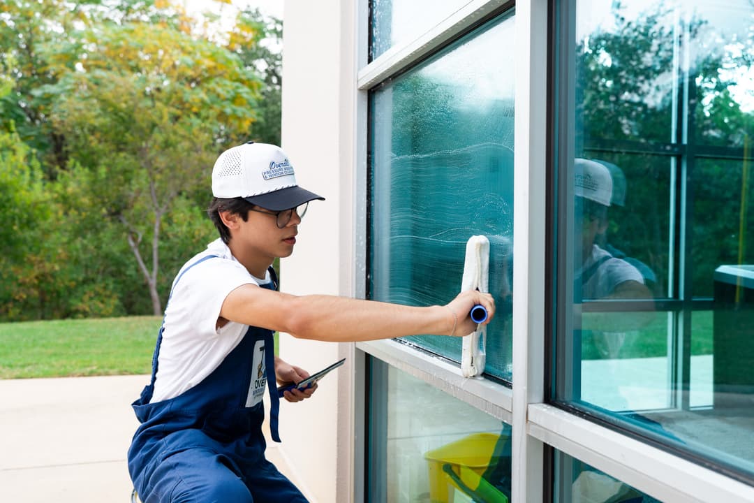 Young worker cleaning a window with a squeegee, wearing a cap and work uniform outdoors.