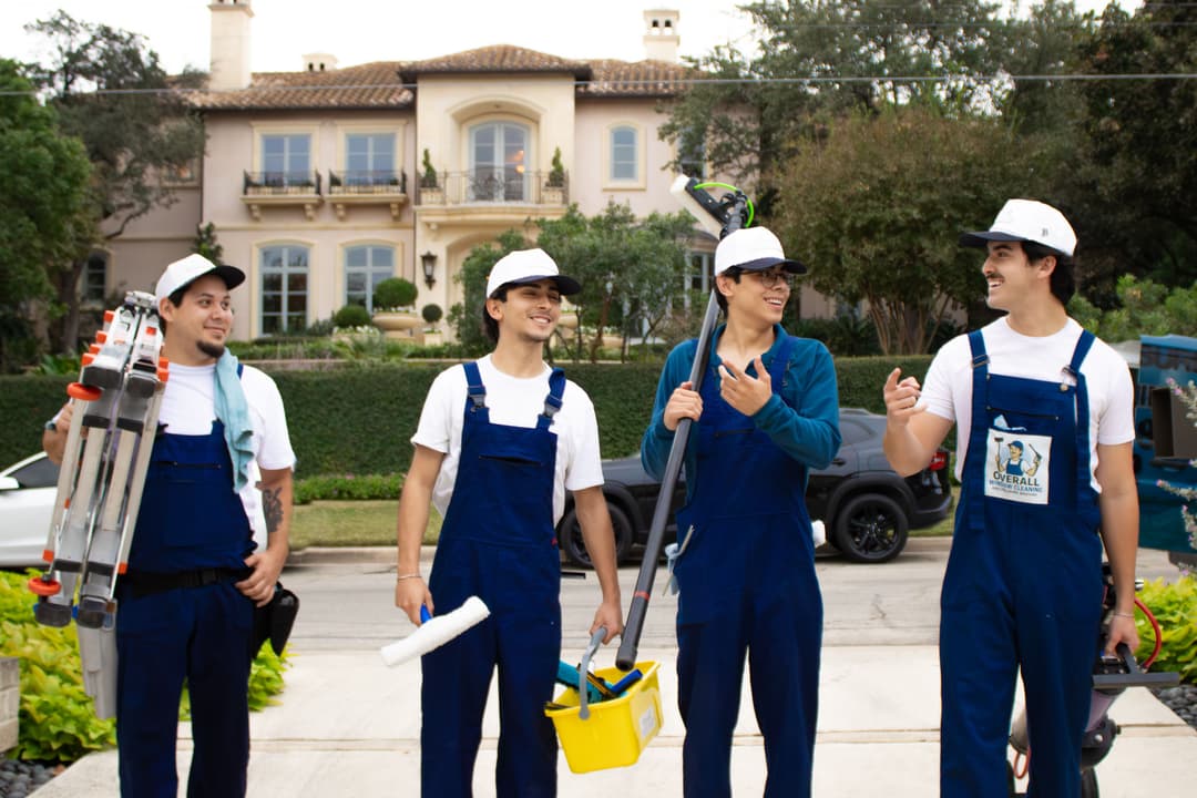 Four workers in blue overalls and hard hats stand on a driveway with cleaning tools and a house behind.