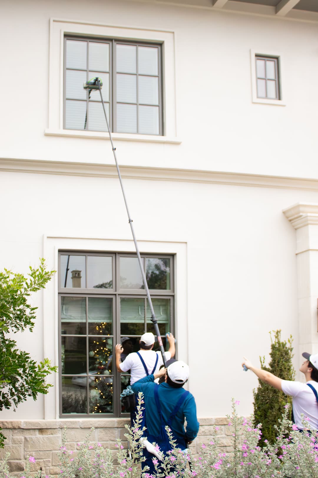Professionals cleaning a tall window using a pole from the ground level of a house.