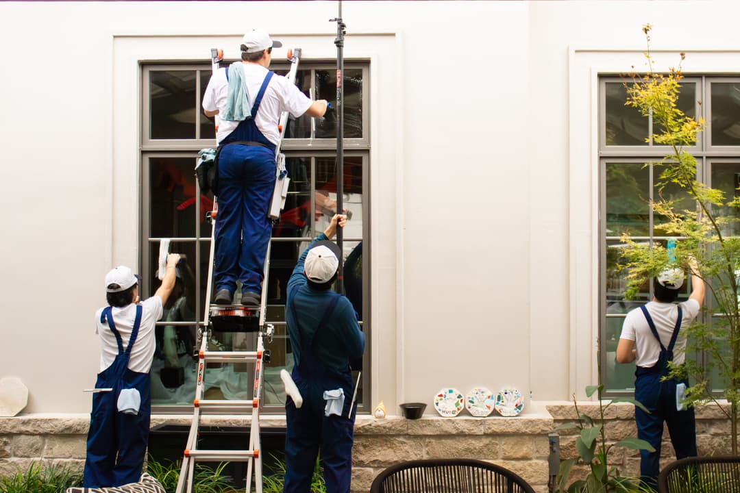 Professional window cleaners in blue uniforms cleaning glass, using ladder and tools.