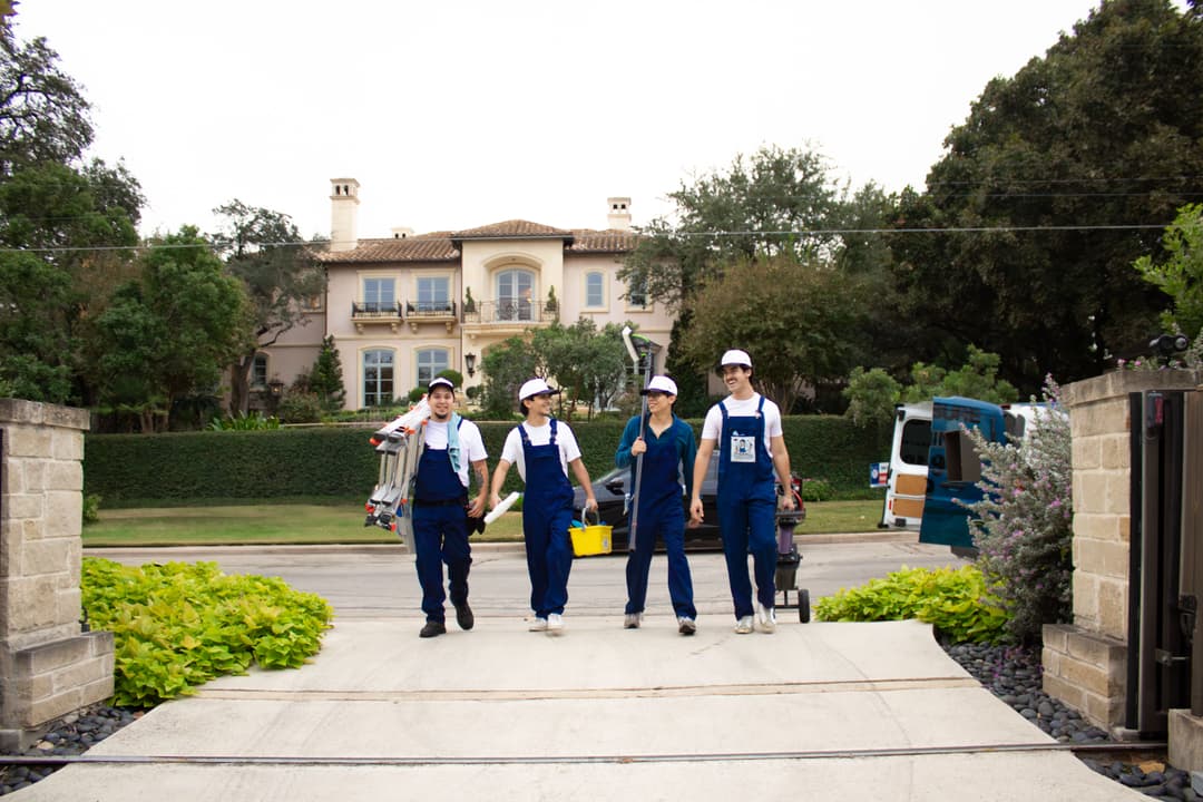 Four service professionals in uniforms walking toward a mansion, carrying tools and equipment.