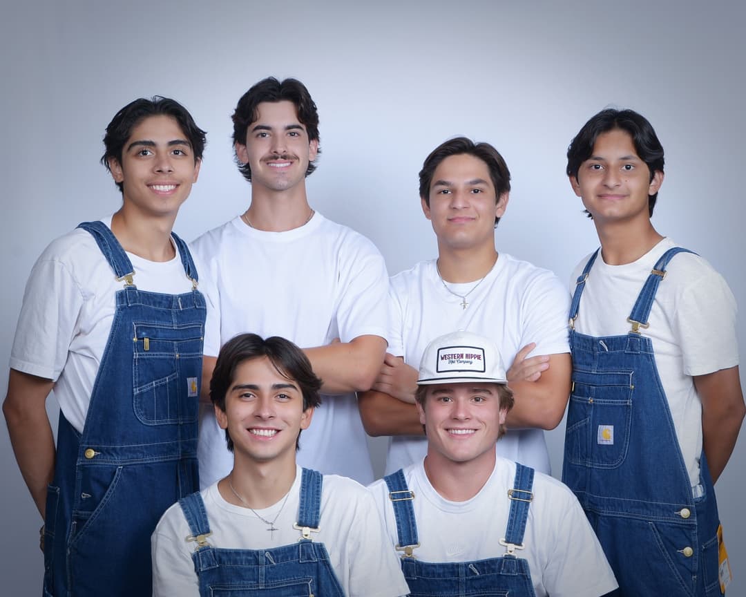 Group of six young men in denim overalls and white shirts posing together.